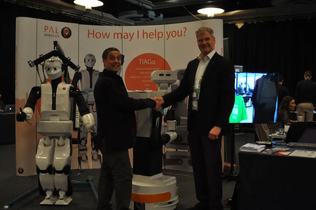 PAL Robotics' CEO Francesco Ferro shaking hands at ERF2017 with TIAGo robot and REEM-C in the background