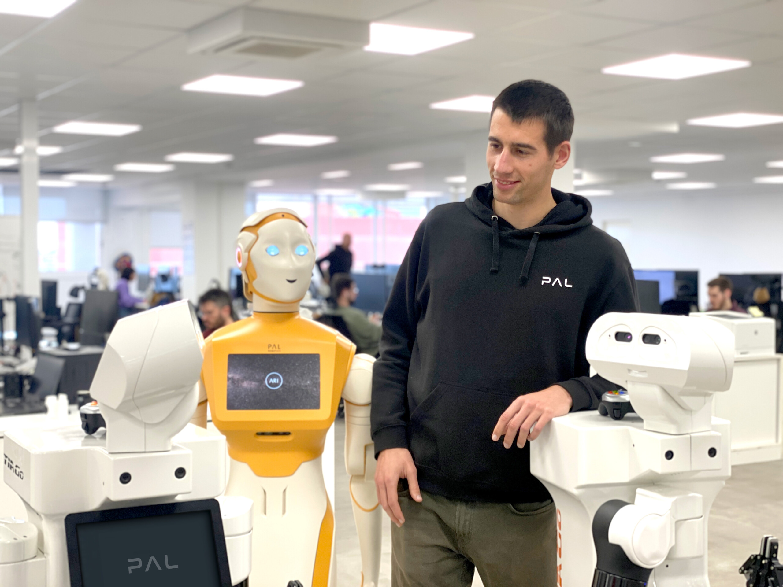 The image depicts Ferran Gebelli interacting with a TIAGo and ARI robots by PAL Robotics. The setting appears to be a busy office or research lab with several individuals at their workstations in the background. The focus is on Ferran and the robot, highlighting a moment of human-robot interaction. 